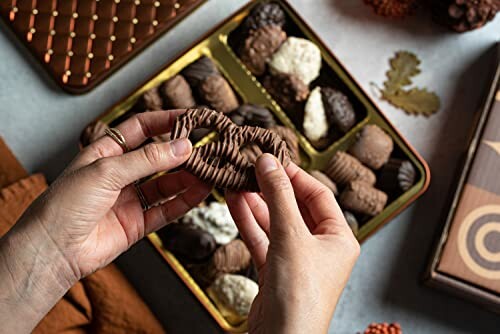 Hands holding a chocolate piece over a box of assorted chocolates.