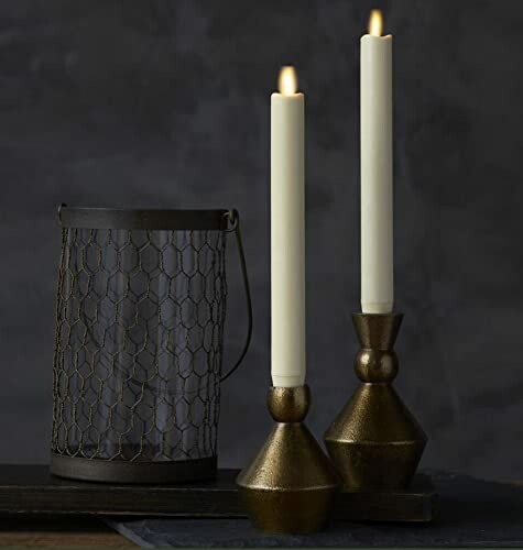 Two candlesticks with lit candles next to a wire mesh lantern.